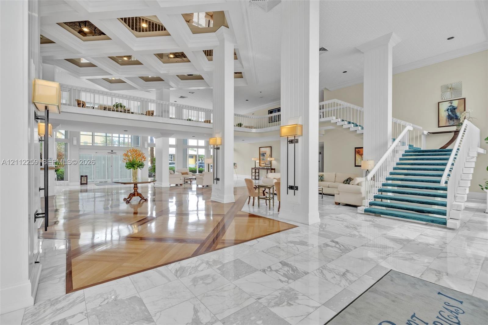 Grand foyer featuring a coffered ceiling, polished stone flooring with wood-finish inlays, structural columns, and an open staircase with a custom railing