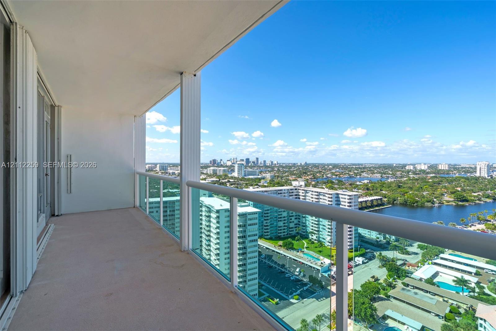 Covered balcony featuring concrete flooring, a glass-panel railing with a metallic handrail, and an expansive cityscape and waterway backdrop Covered balcony featuring concrete flooring, a glass-panel railing with a metallic handrail, and an expansive cityscape and waterway backdrop