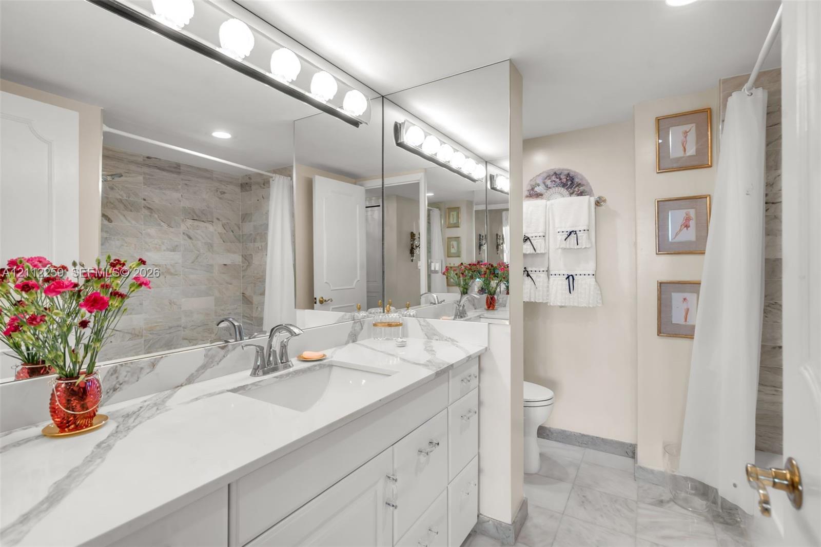 Bathroom featuring a white vanity with stone-patterned countertop, integrated basin, full-width mirror, multi-bulb light fixture, and stone-tiled shower surround