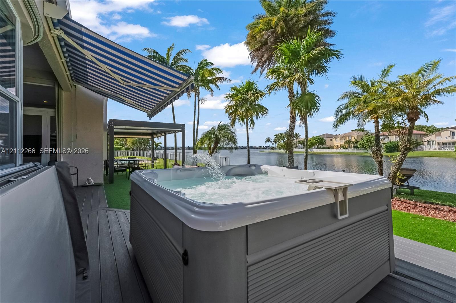 Outdoor spa on a composite deck featuring a retractable striped awning, overhead pergola, and water frontage