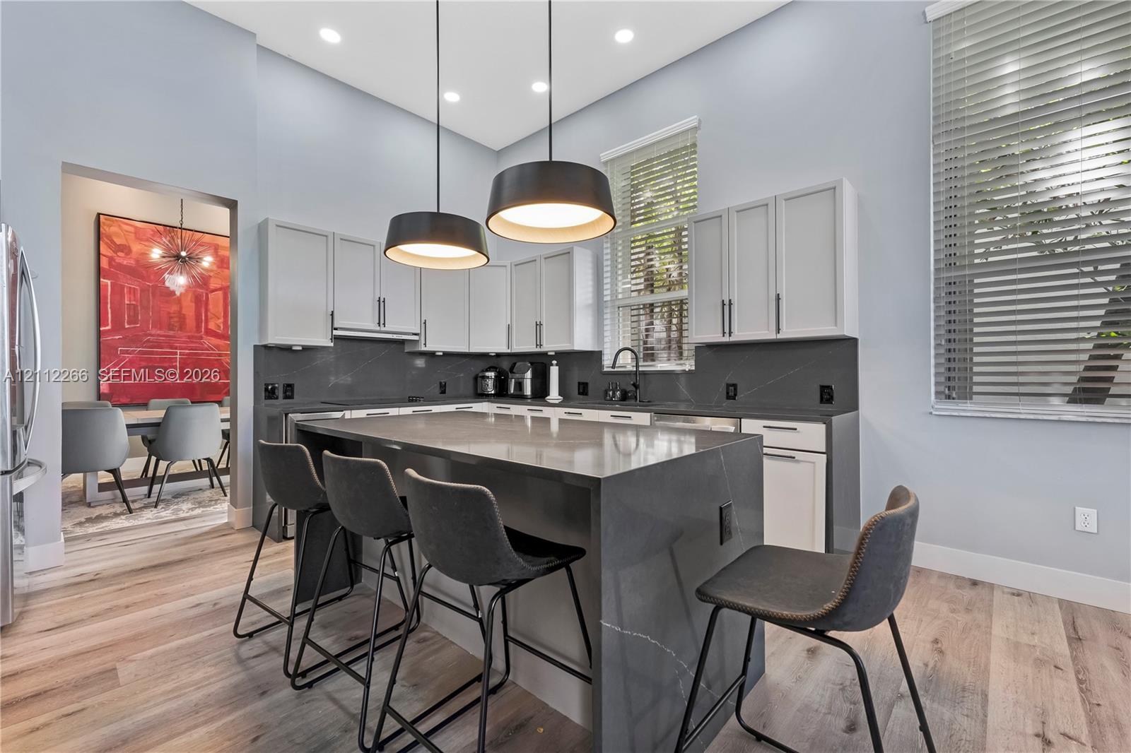 Kitchen featuring a central island with a solid-surface waterfall countertop, white cabinetry with dark hardware, dark solid-surface backsplash, wood-finish flooring, and dual pendant lighting Kitchen featuring a central island with a solid-surface waterfall countertop, white cabinetry with dark hardware, dark solid-surface backsplash, wood-finish flooring, and dual pendant lighting