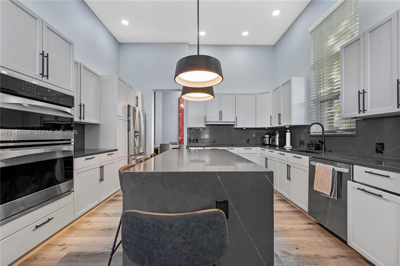 Kitchen featuring white shaker-style cabinetry, dark stone countertops with a waterfall-edge island, wood-finish flooring, dual wall ovens, and matte black hardware Kitchen featuring white shaker-style cabinetry, dark stone countertops with a waterfall-edge island, wood-finish flooring, dual wall ovens, and matte black hardware