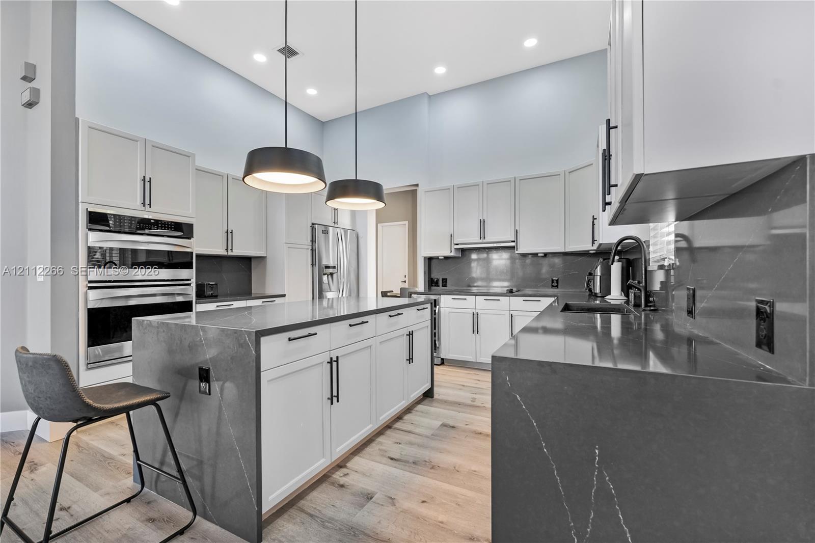 Kitchen featuring a waterfall-edge island, dark stone-finish countertops, ceiling-height cabinetry, double wall ovens, and wood-finish flooring Kitchen featuring a waterfall-edge island, dark stone-finish countertops, ceiling-height cabinetry, double wall ovens, and wood-finish flooring
