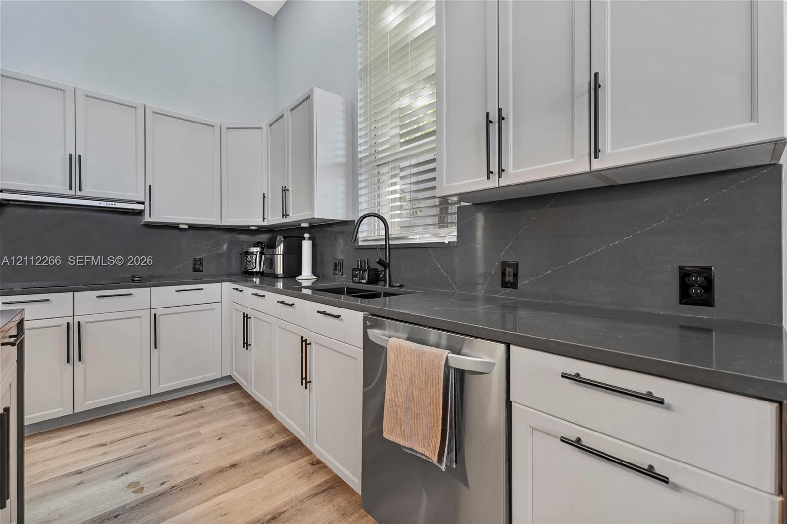 Kitchen featuring white shaker-style cabinetry, dark stone countertops, stainless steel dishwasher, matte black hardware, and wood-finish flooring Kitchen featuring white shaker-style cabinetry, dark stone countertops, stainless steel dishwasher, matte black hardware, and wood-finish flooring
