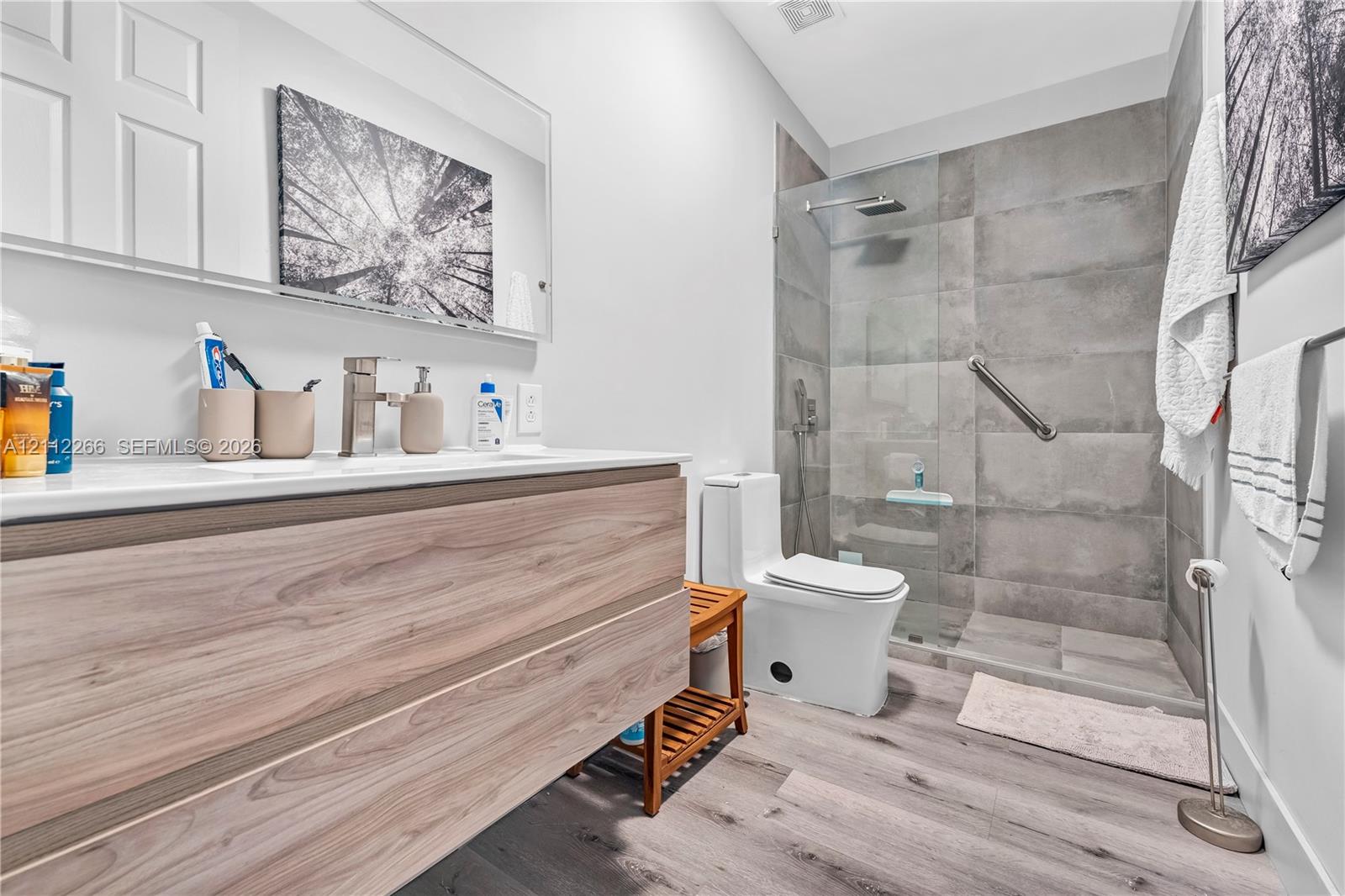 Floating wood-finish vanity with a clean-lined countertop, walk-in shower featuring large-format grey wall tile and a glass partition, rainfall showerhead, and wood-finish flooring