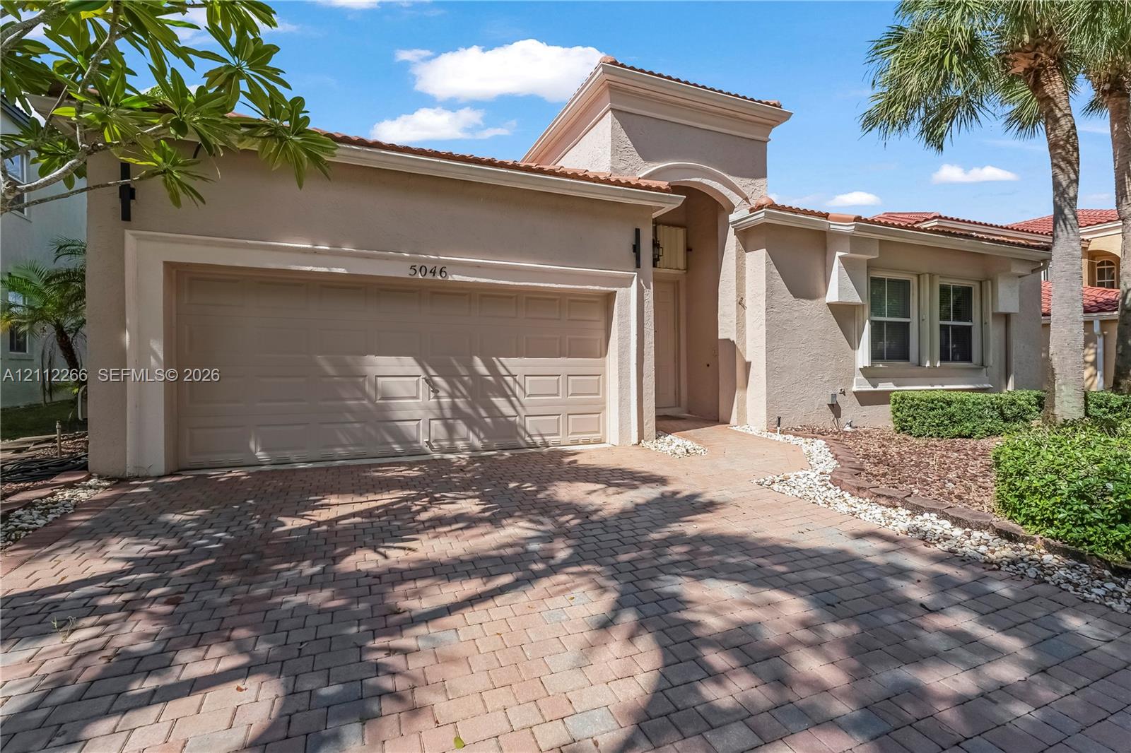 Stucco exterior with a paved brick driveway, decorative roofline trim, arched entry, and integrated multi-panel garage door