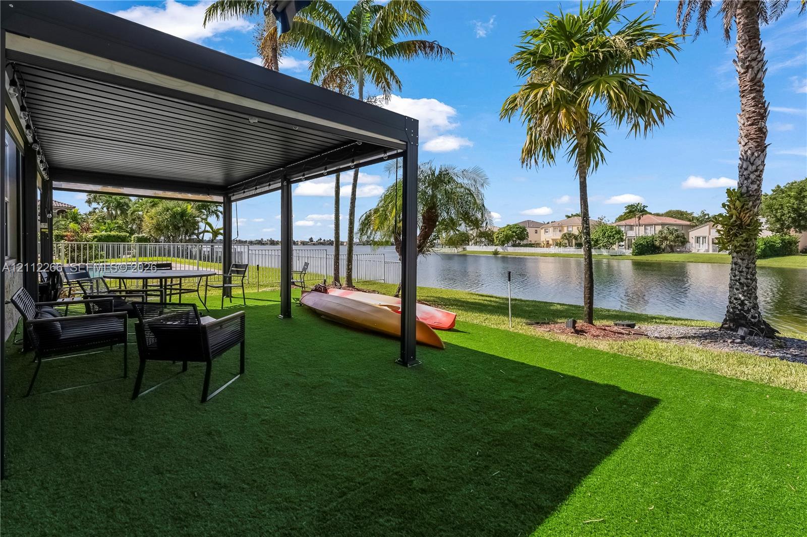 Covered patio featuring a dark metal pergola, synthetic grass lawn, perimeter fencing, and water frontage with mature palm trees