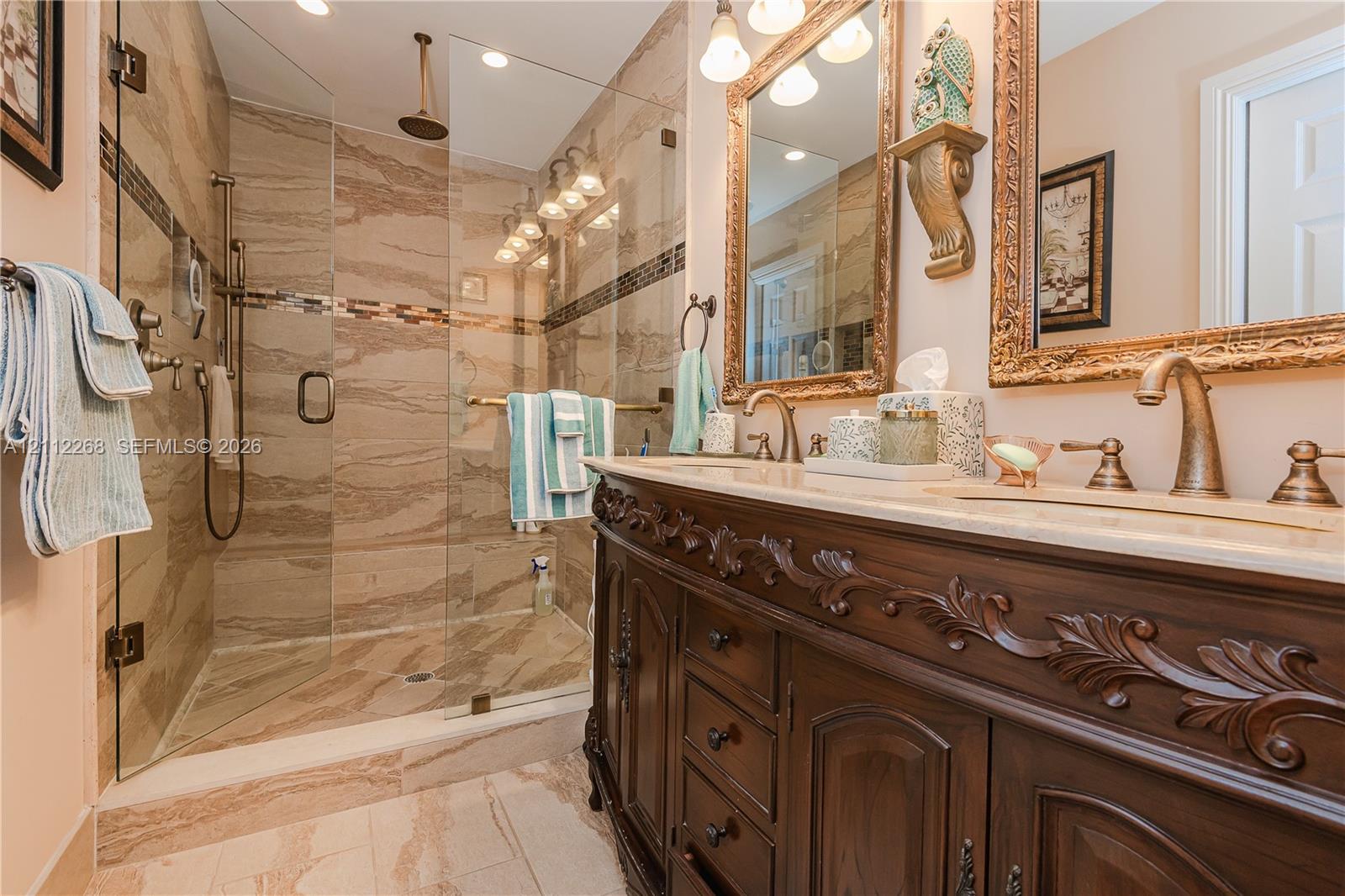 Bathroom featuring an expansive wood vanity with intricate carved detailing, a glass-enclosed walk-in shower with floor-to-ceiling tile surround, stone-finish countertops, dual-sink configuration, and brass-tone plumbing fixtures