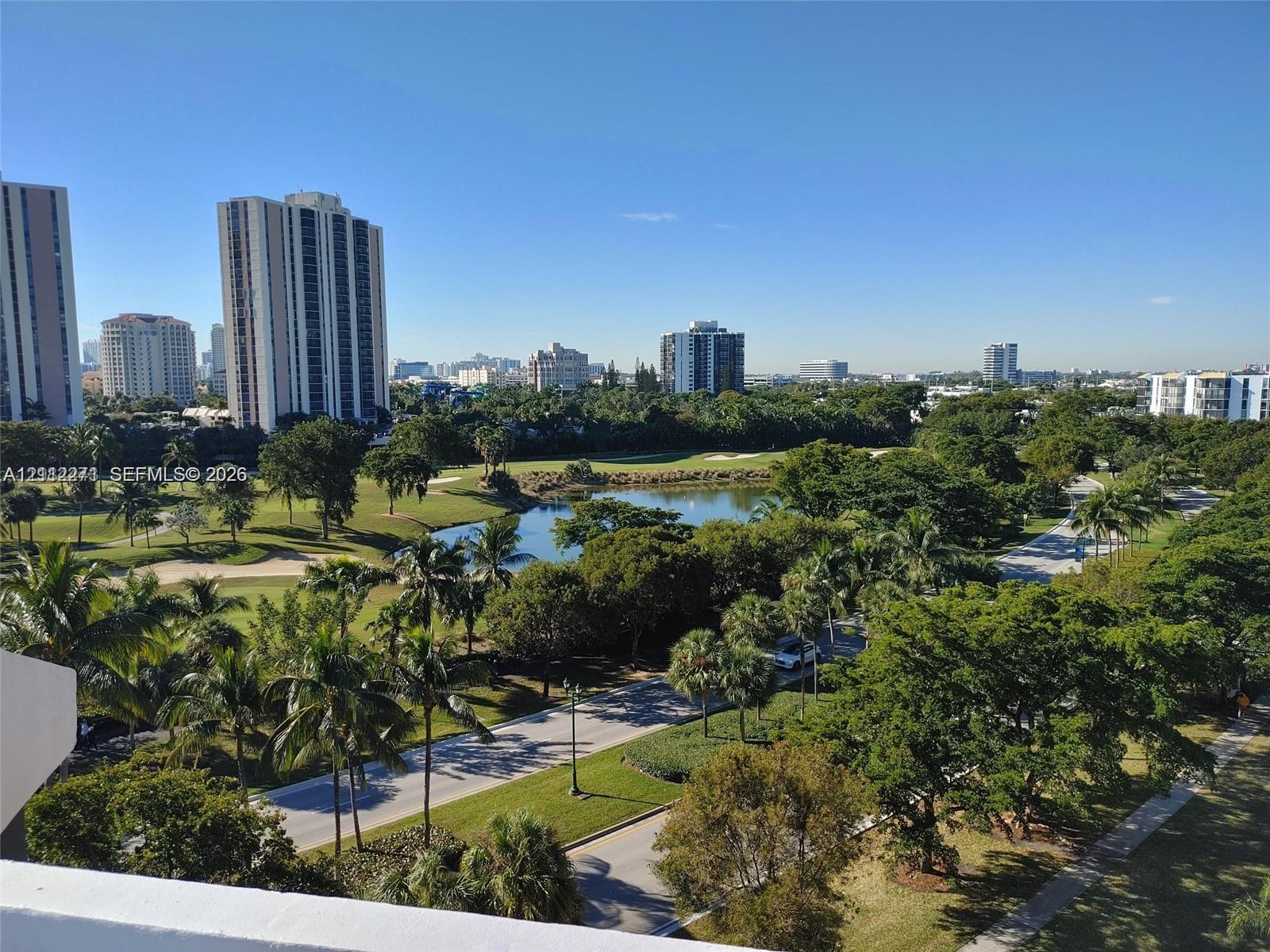 Elevated balcony perspective featuring expansive scenery of a manicured golf course, a central water feature, mature trees, and high-rise urban architecture