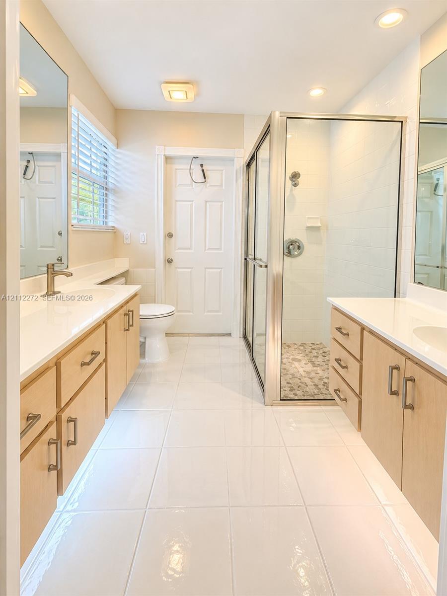 Dual-vanity bathroom featuring light wood cabinetry, quartz countertops, tile flooring, and a glass-enclosed shower with pebble-stone tiling