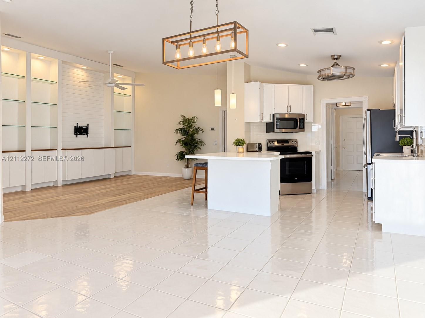 Open-concept interior featuring large-format tile flooring, wood-finish flooring, custom built-in cabinetry with glass shelving, a center island with quartz countertops, and a linear metal pendant light fixture Open-concept interior featuring large-format tile flooring, wood-finish flooring, custom built-in cabinetry with glass shelving, a center island with quartz countertops, and a linear metal pendant light fixture