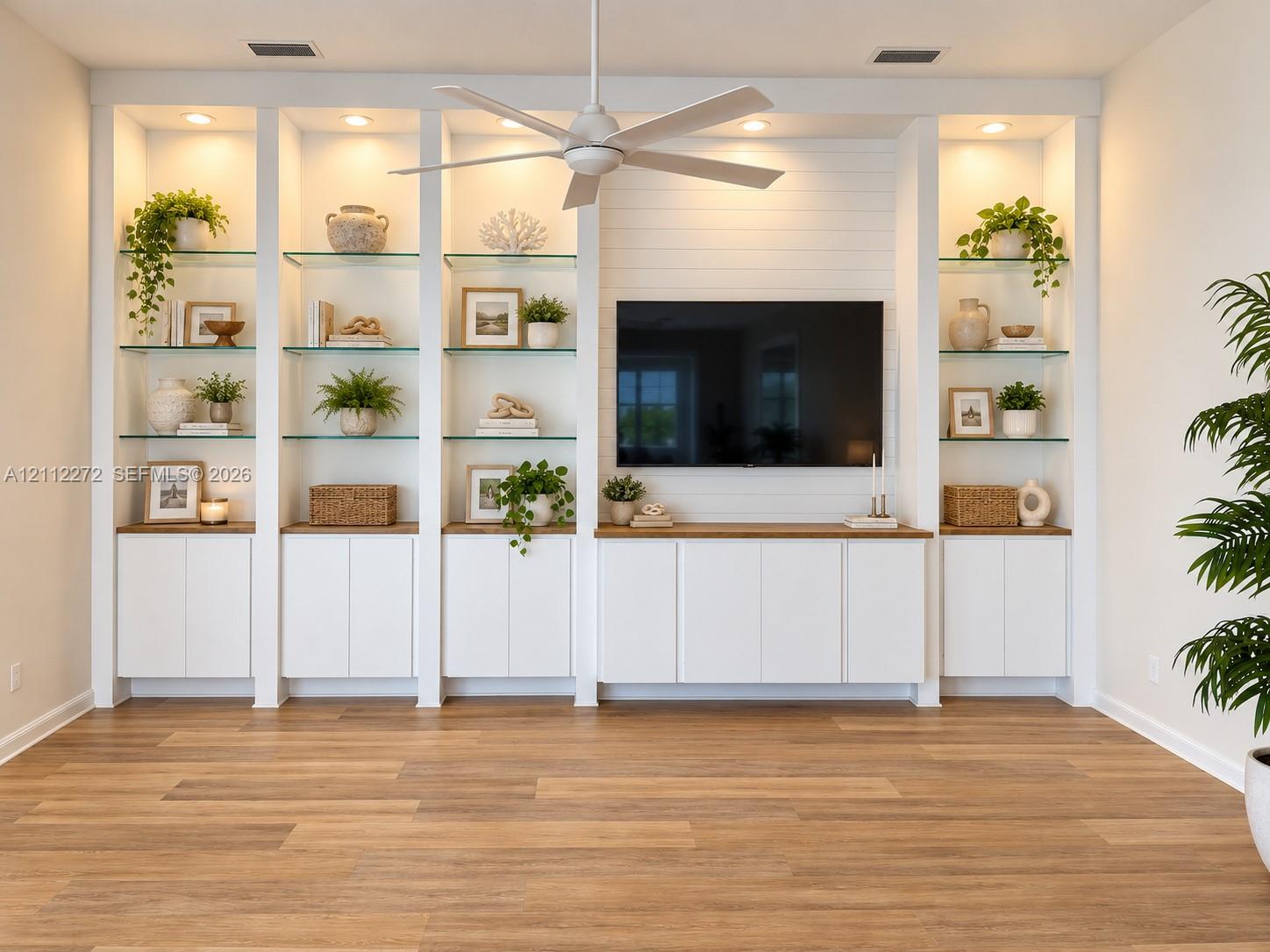 Custom floor-to-ceiling built-in cabinetry featuring white shaker-style doors, wood-finish countertops, glass shelving, and integrated recessed lighting, accented by a white shiplap wall and a matching ceiling fan