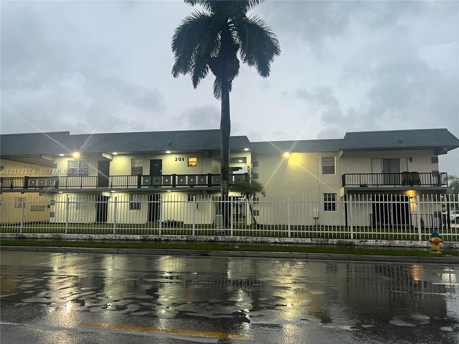 Multi-level exterior featuring a light-colored stucco facade, shingled roofing, second-story railed balconies, and perimeter metal fencing Multi-level exterior featuring a light-colored stucco facade, shingled roofing, second-story railed balconies, and perimeter metal fencing