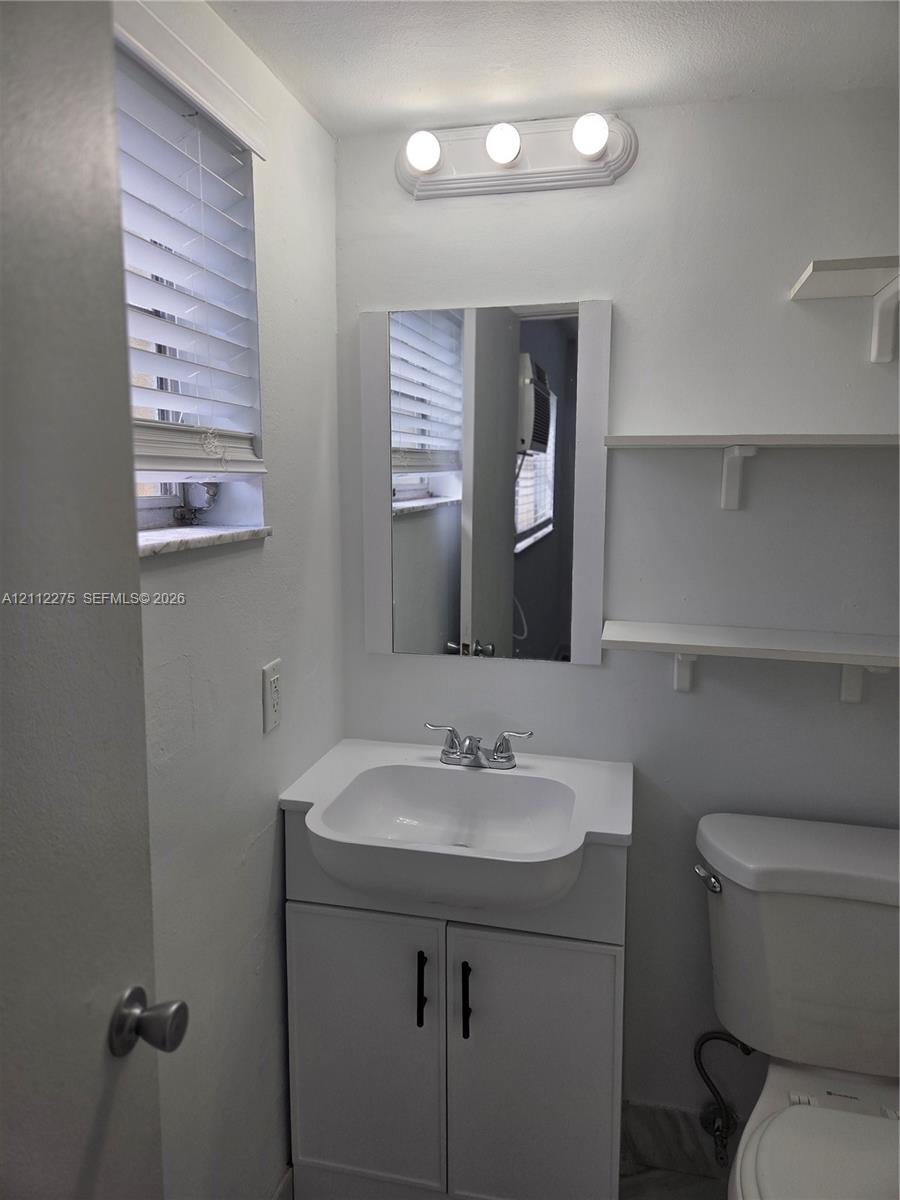 White vanity with a rectangular sink, chrome dual-handle faucet, frameless mirror, three-light fixture, and wall-mounted shelving