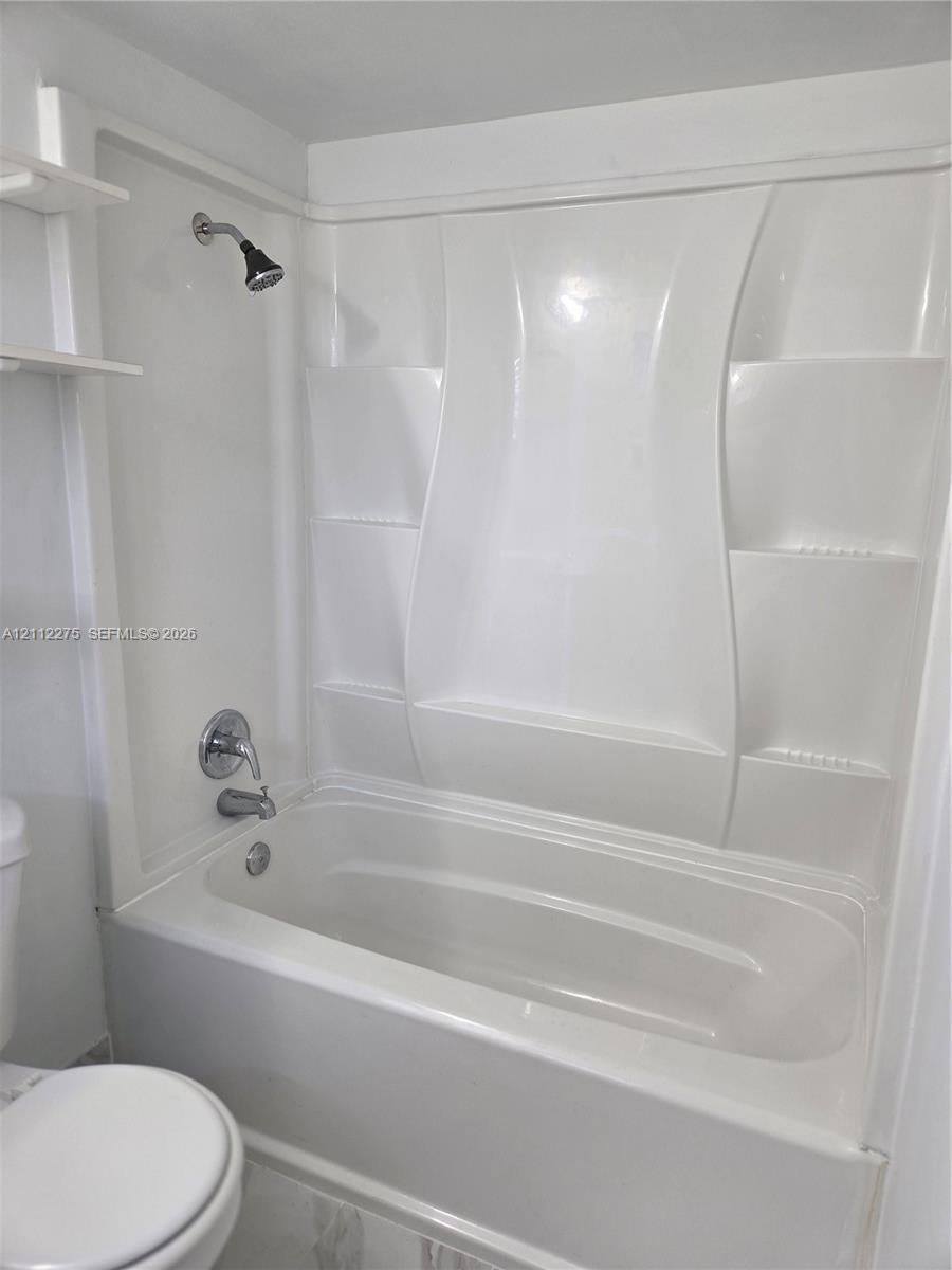 White acrylic bathtub and shower surround featuring integrated shelving, chrome fixtures, and a wall-mounted shower head