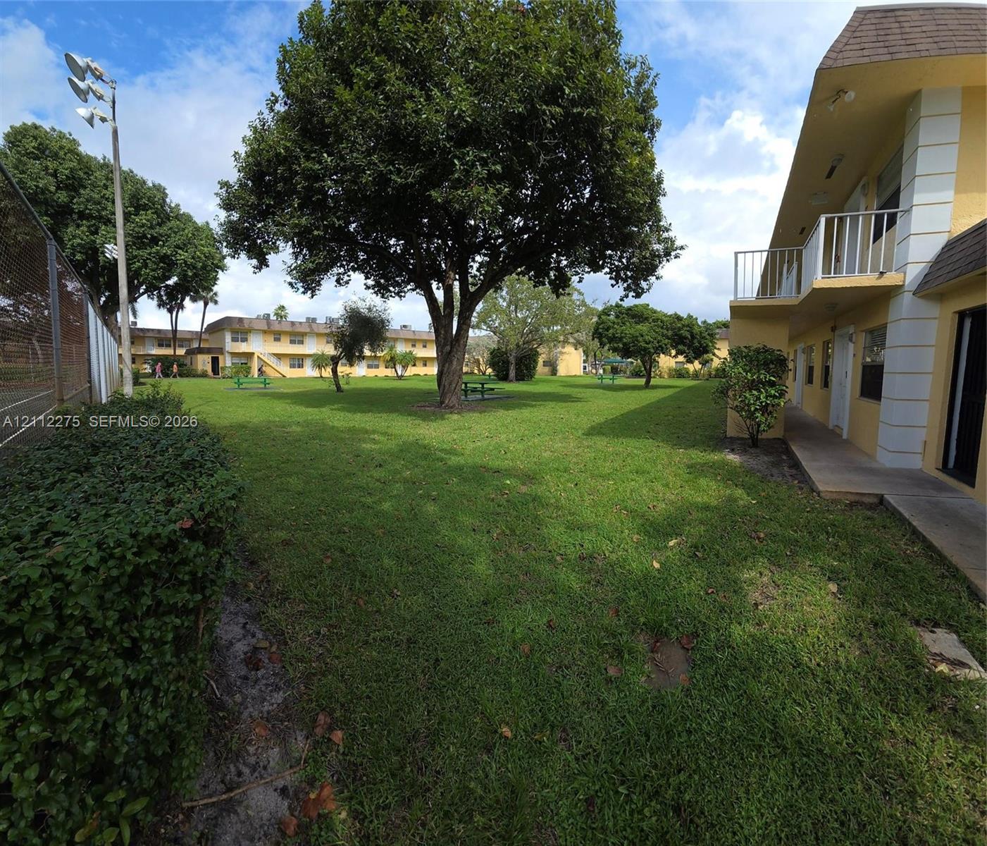 Expansive common area featuring a manicured lawn, mature trees, metal perimeter fencing, concrete walkways, and elevated exterior balconies