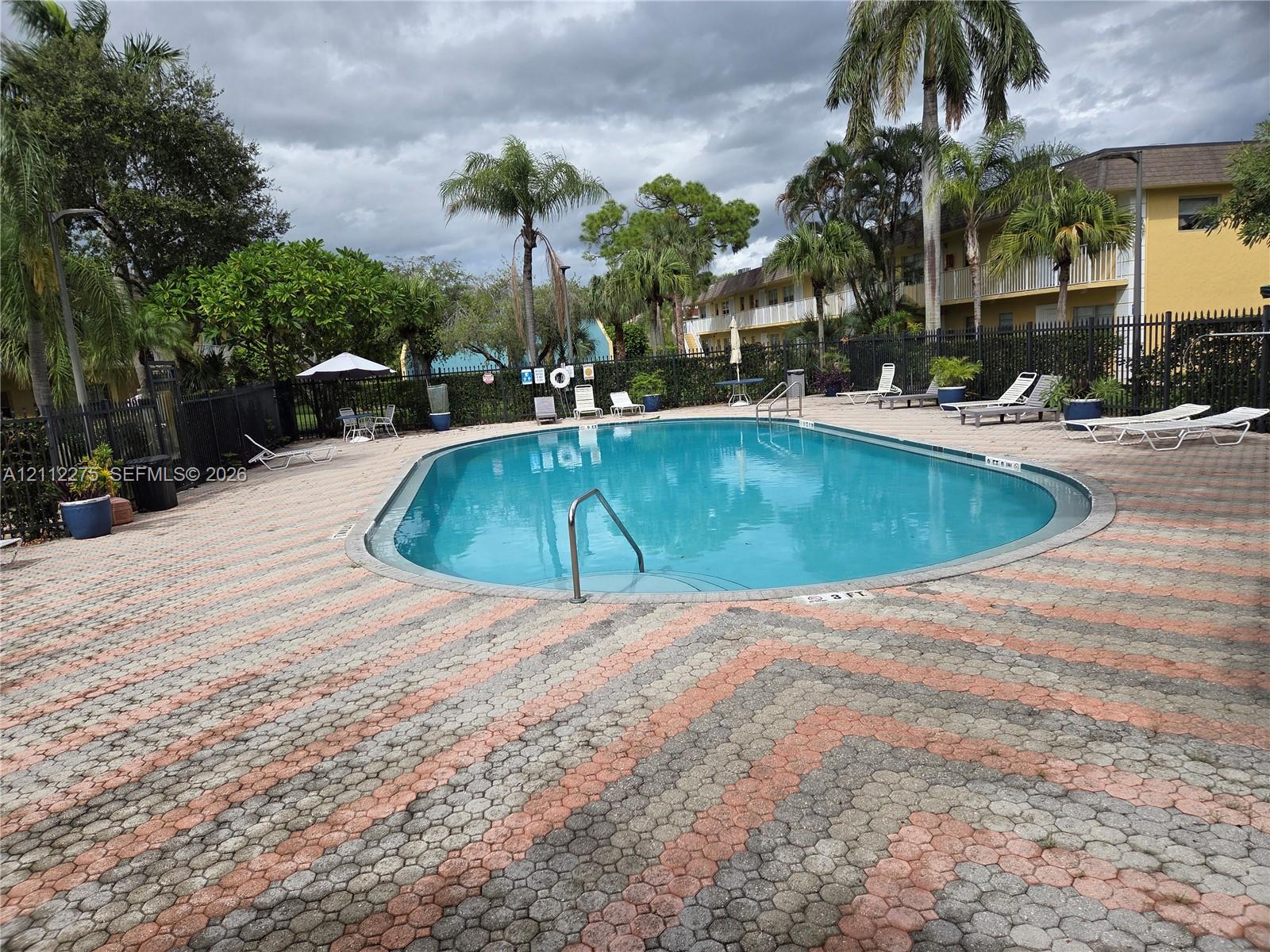 Oval swimming pool featuring a metal entry rail, surrounded by patterned paver hardscaping and perimeter fencing