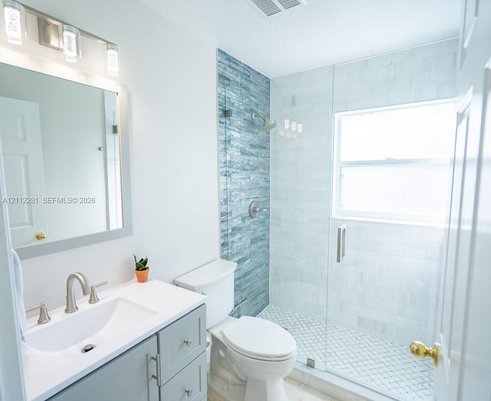 Bathroom featuring a vanity with a white countertop and rectangular undermount sink, a walk-in shower with glass enclosure, textured blue tile accent wall, and three-light brushed-nickel fixture above the mirror