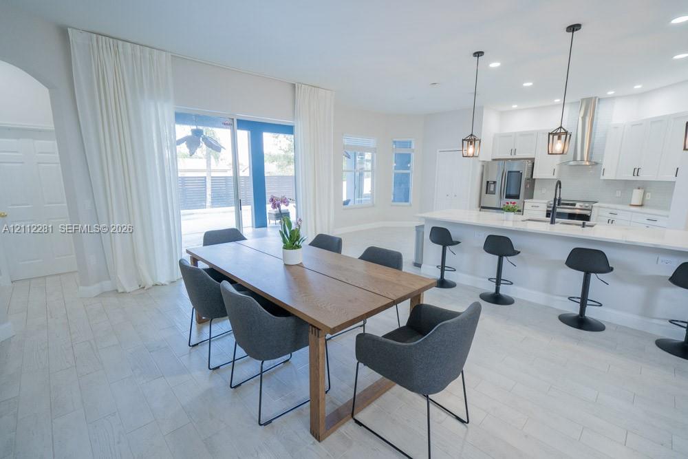 Open-concept kitchen featuring white shaker-style cabinetry, quartz countertops, a stainless steel range hood, pendant lighting, and wood-finish tile flooring