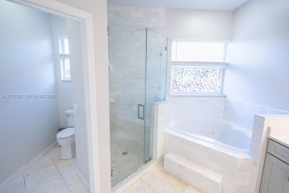 Walk-in glass-enclosed shower featuring stone-tile surround, oversized soaking tub with tile deck, light-finish tile flooring, and integrated commode area