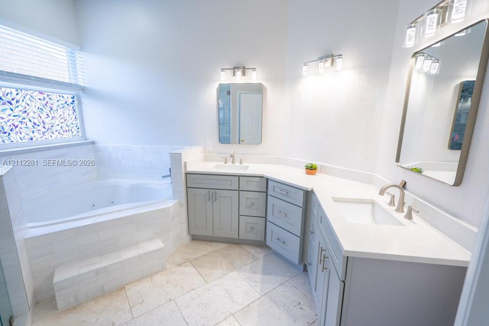 Bathroom featuring a corner soaking tub with a tiled surround, dual-sink vanity with light-colored cabinetry, stone-finish flooring, and wall-mounted vanity lighting