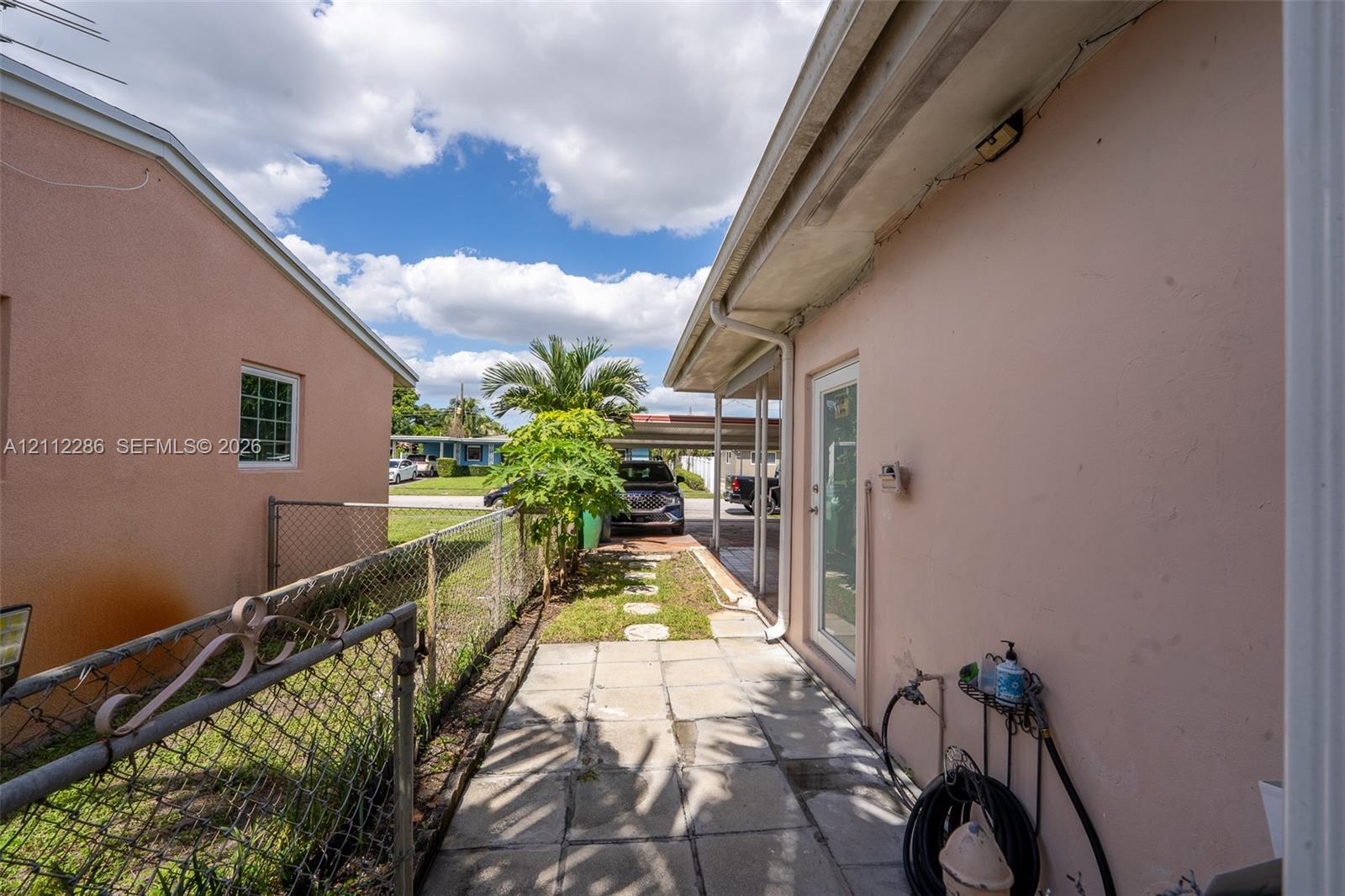 Exterior side area featuring a concrete paver walkway, chain-link fencing, a grass strip with stone pavers, and a metal-roofed carport structure Exterior side area featuring a concrete paver walkway, chain-link fencing, a grass strip with stone pavers, and a metal-roofed carport structure