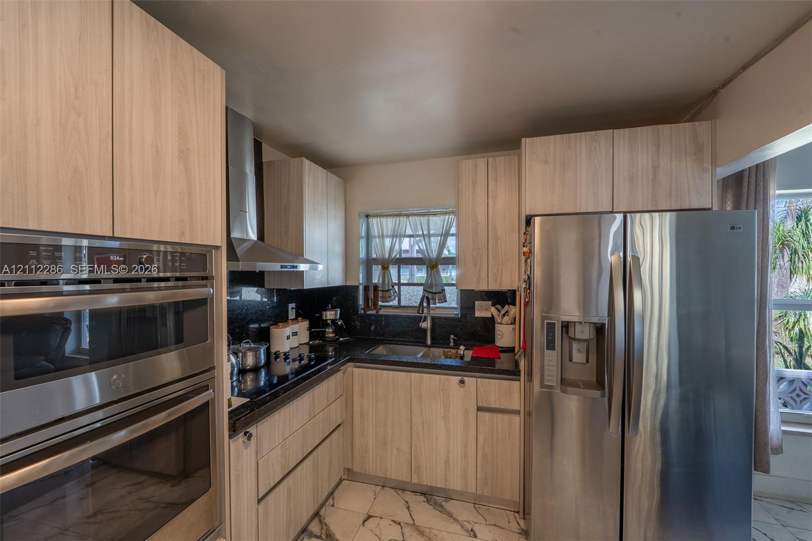 Kitchen featuring light-tone flat-panel cabinetry, dark stone countertops, stainless steel wall oven, stainless steel chimney-style range hood, and large-format tile flooring