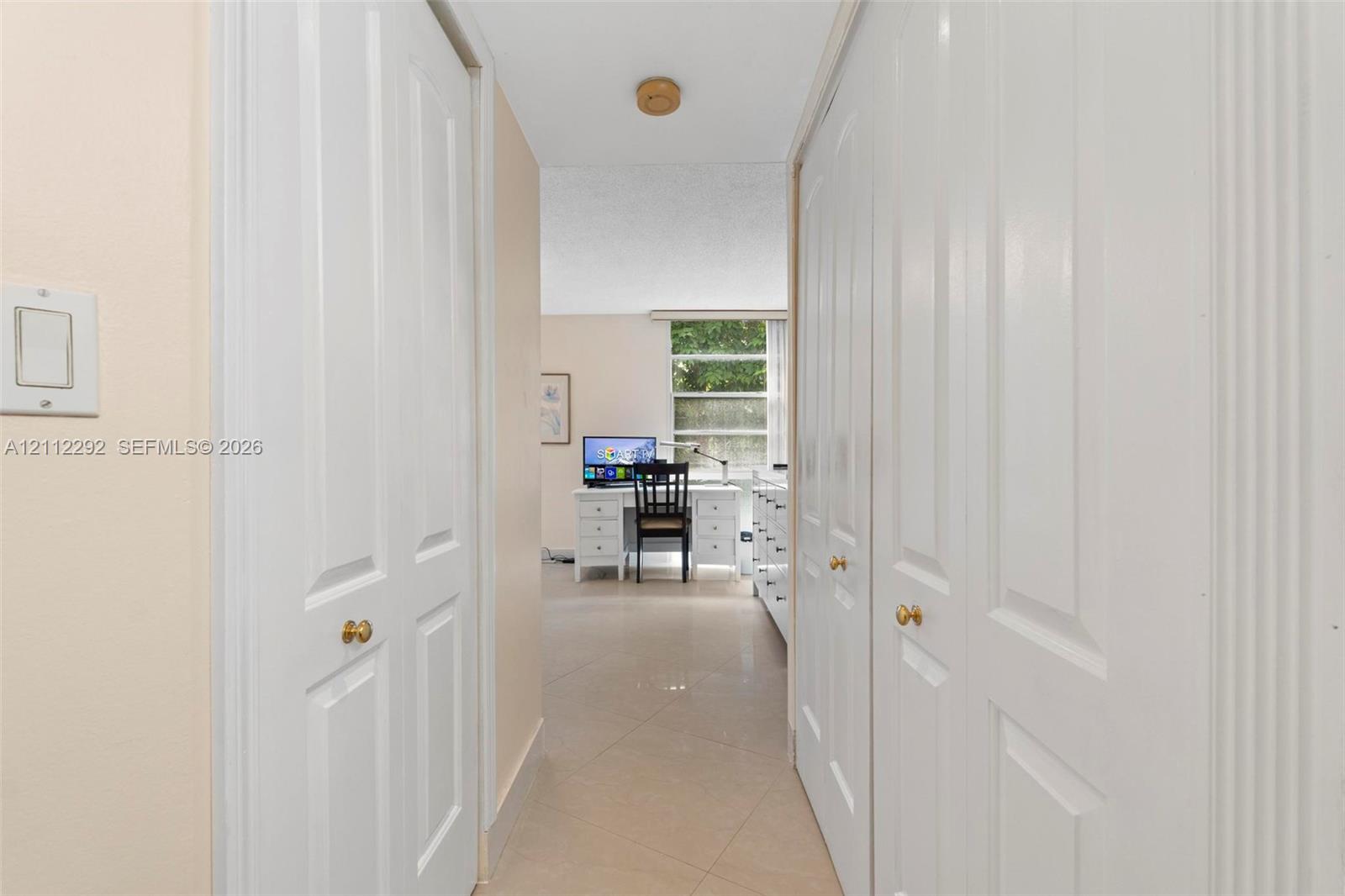 Hallway featuring dual bifold closets with brass-finish hardware and neutral tile flooring