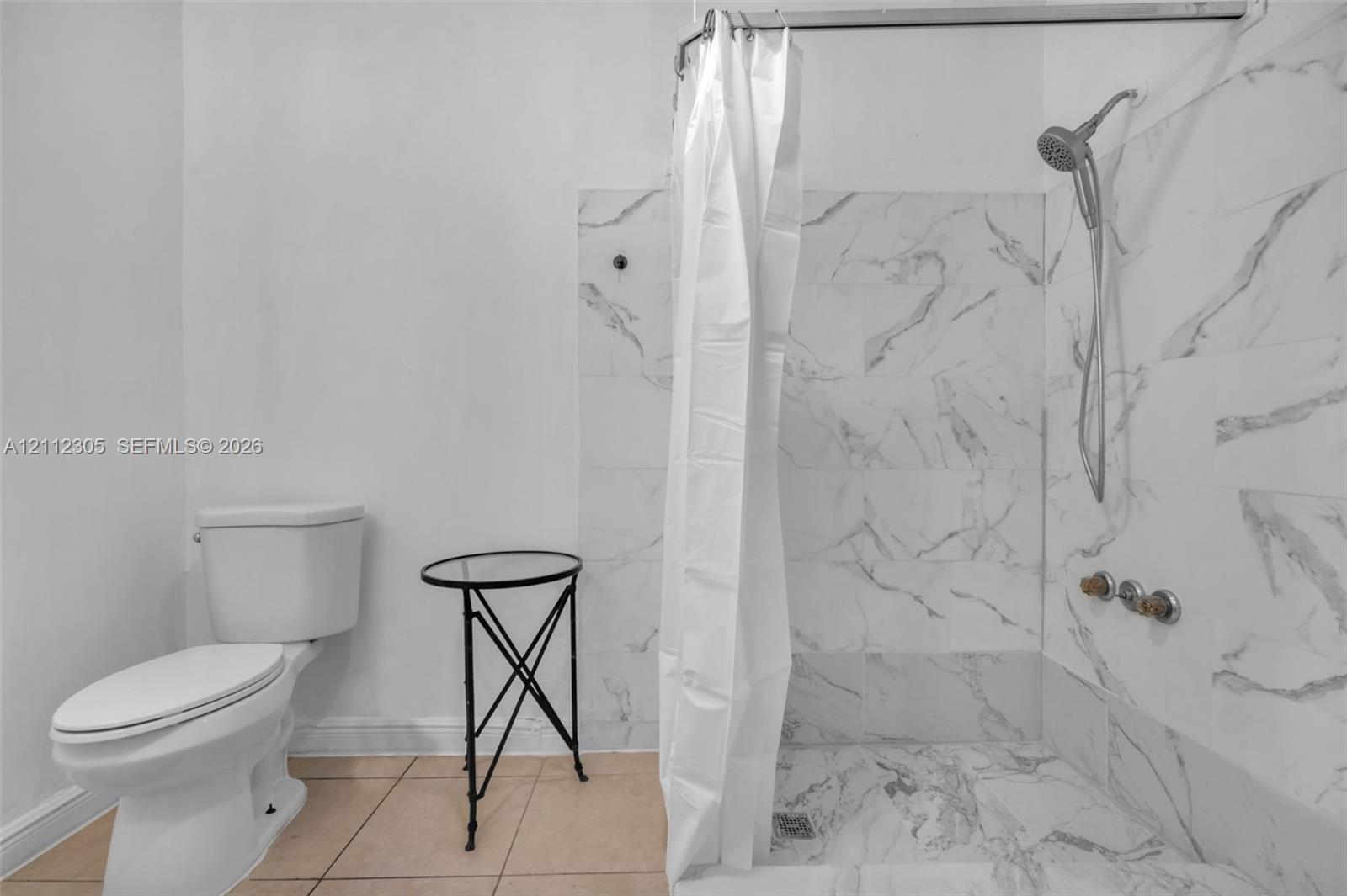 Walk-in shower enclosure featuring large-format wall-to-floor veined tile, handheld shower fixture, and ceramic toilet