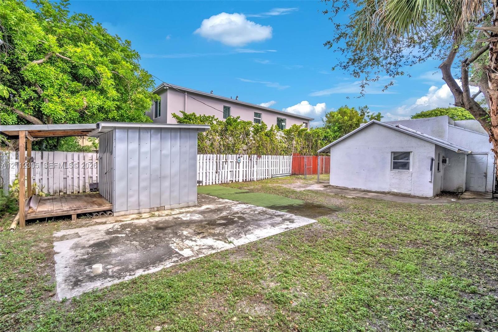 Fenced backyard featuring an auxiliary structure with a covered porch, an open-air concrete pad, and mature trees