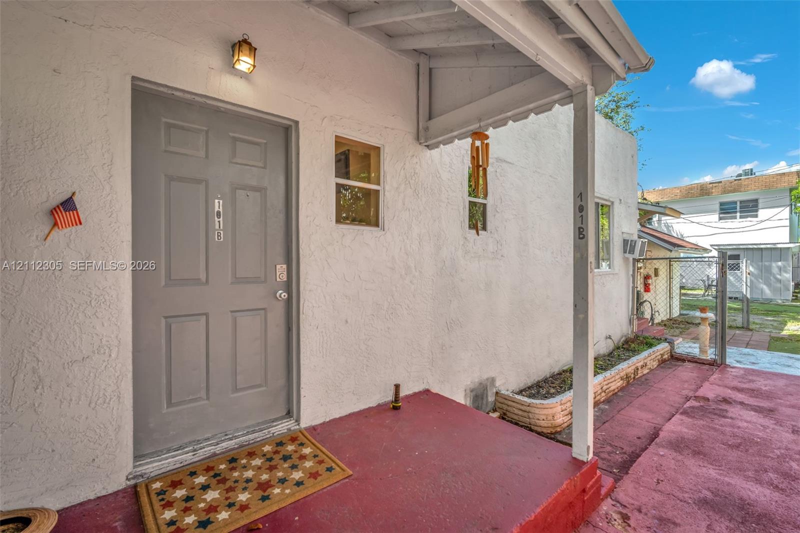 Covered entryway featuring a textured stucco exterior, a solid six-panel door, an exterior wall-mounted light fixture, and a masonry planter box