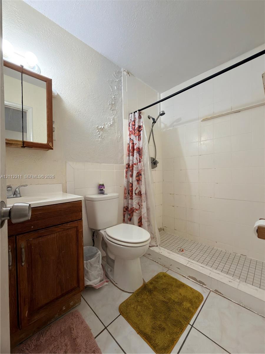 Bathroom featuring a wood-finish vanity with a single-basin sink, wall-mounted medicine cabinet, white tile wall surround, tile shower flooring, and ceramic floor tile