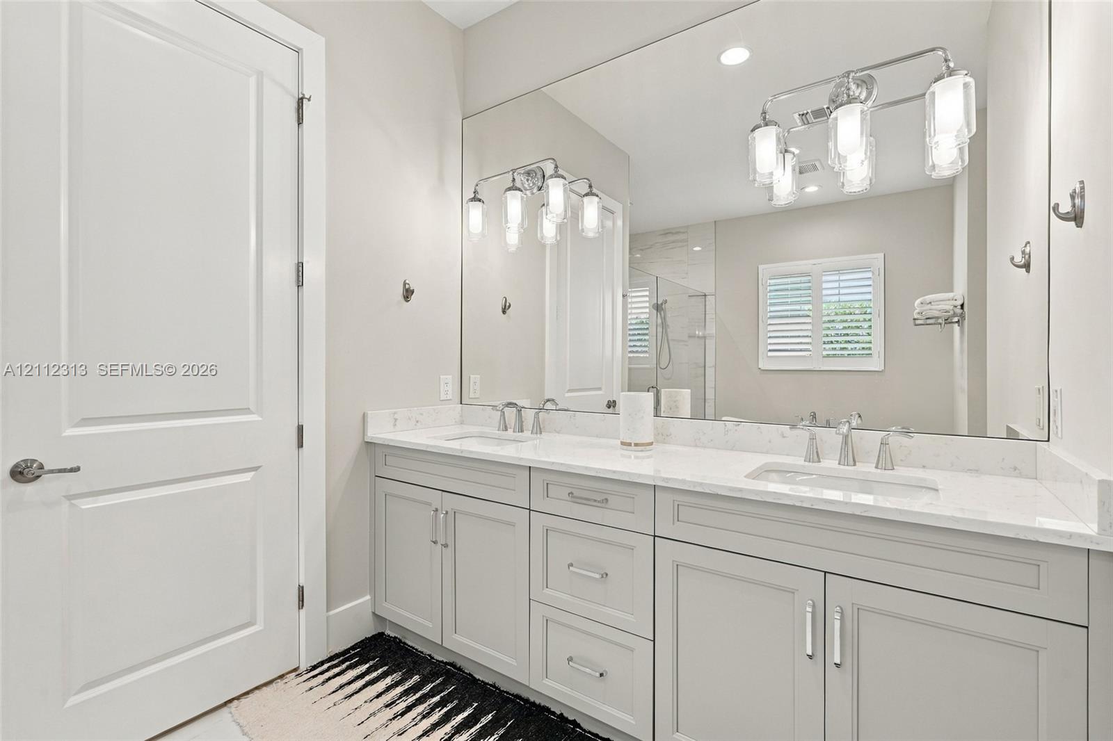 Double vanity featuring light-toned cabinetry, stone-finish countertops, undermount sinks, polished metal fixtures, and a wall-to-wall mirror with multi-bulb integrated lighting