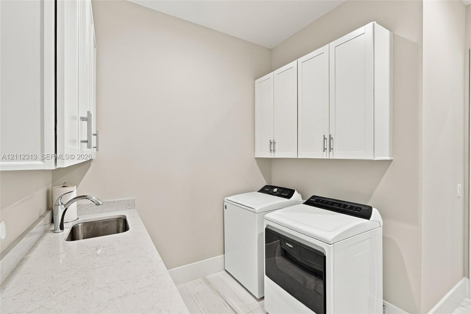 Laundry area featuring white shaker-style wall cabinets, a light-toned countertop, an integrated stainless steel sink with a chrome faucet, and wood-finish flooring