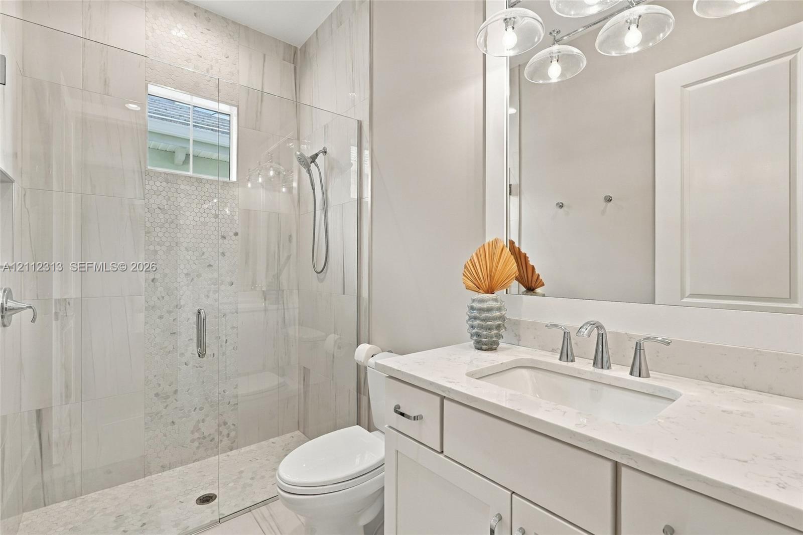 Bathroom featuring a vanity with a white countertop and undermount sink, a walk-in shower with a glass enclosure, floor-to-ceiling tilework, and a multi-bulb light fixture