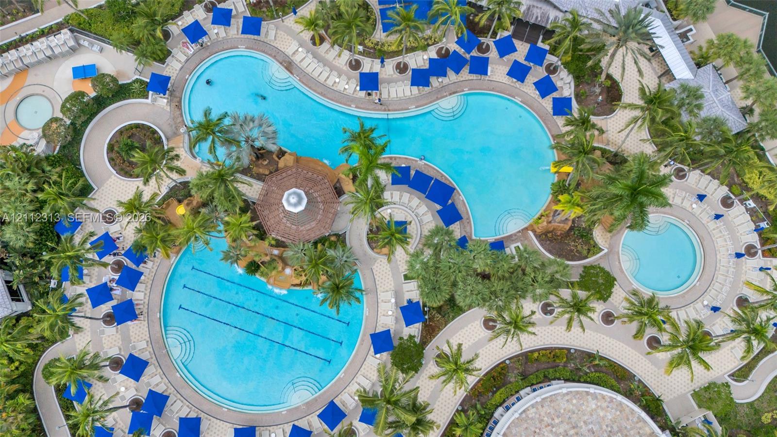 Aerial view of a sprawling pool complex featuring multiple swimming pools, a dedicated lap lane section, a central gazebo, and expansive paved decking surrounded by palm trees