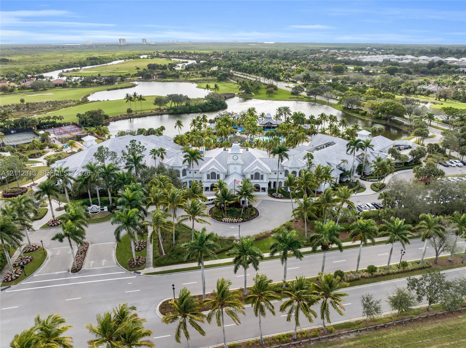 Aerial perspective of a multi-winged facility featuring a central clock tower, expansive pond frontage, mature palm tree landscaping, and paved circular drive