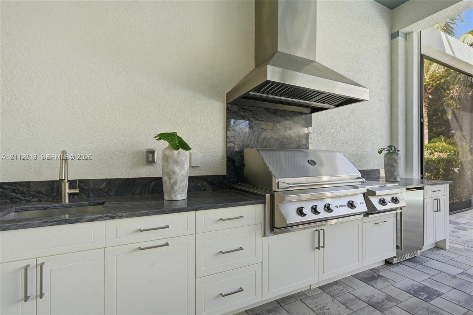 Outdoor kitchen featuring white shaker-style cabinetry, dark natural stone countertops and backsplash, stainless steel built-in grill, commercial-grade ventilation hood, and under-mount sink with modern faucet