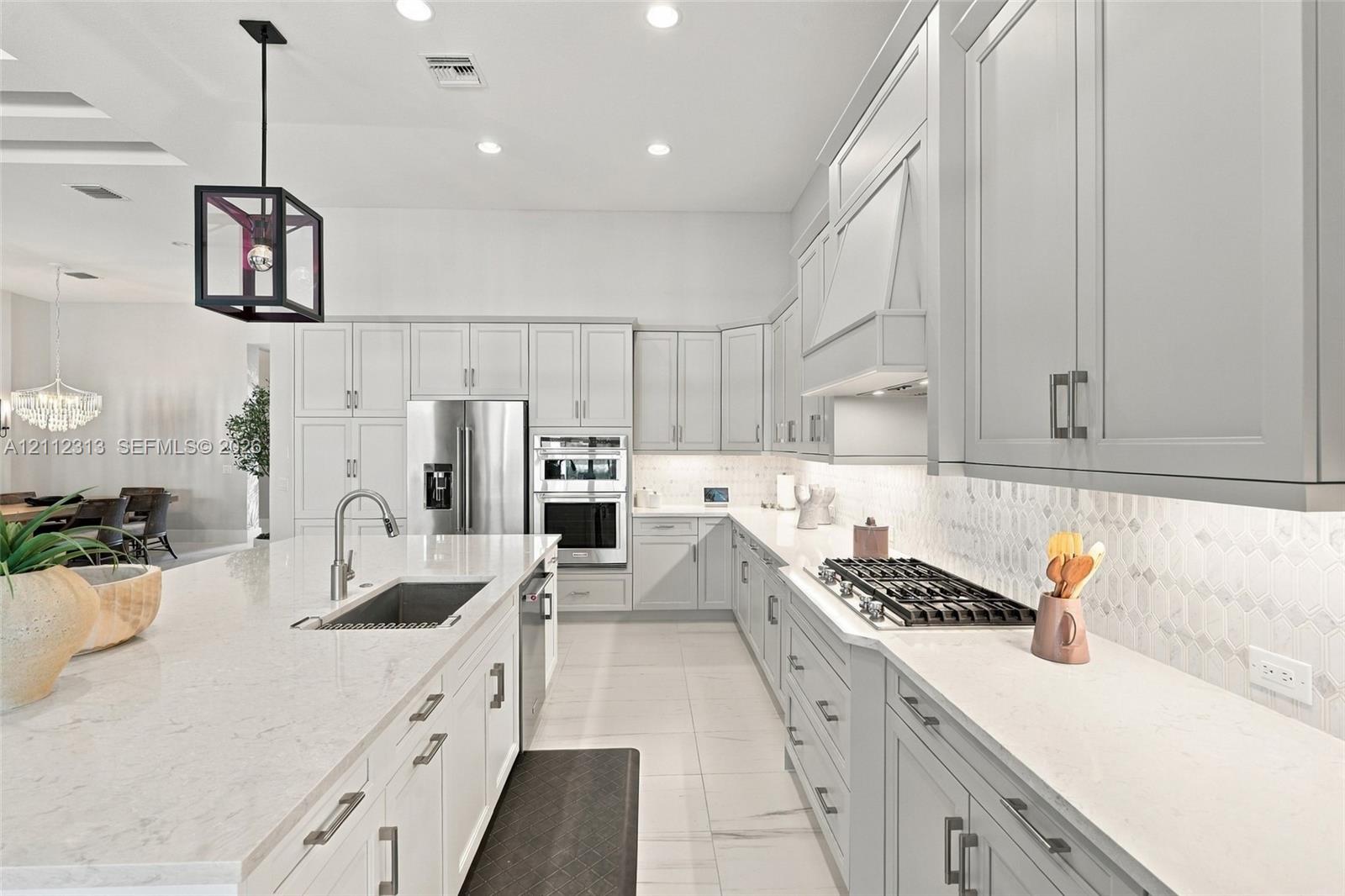 Kitchen featuring light-toned cabinetry, a spacious island with a built-in sink, stone-finish countertops, stainless steel appliances, and a geometric pendant light fixture