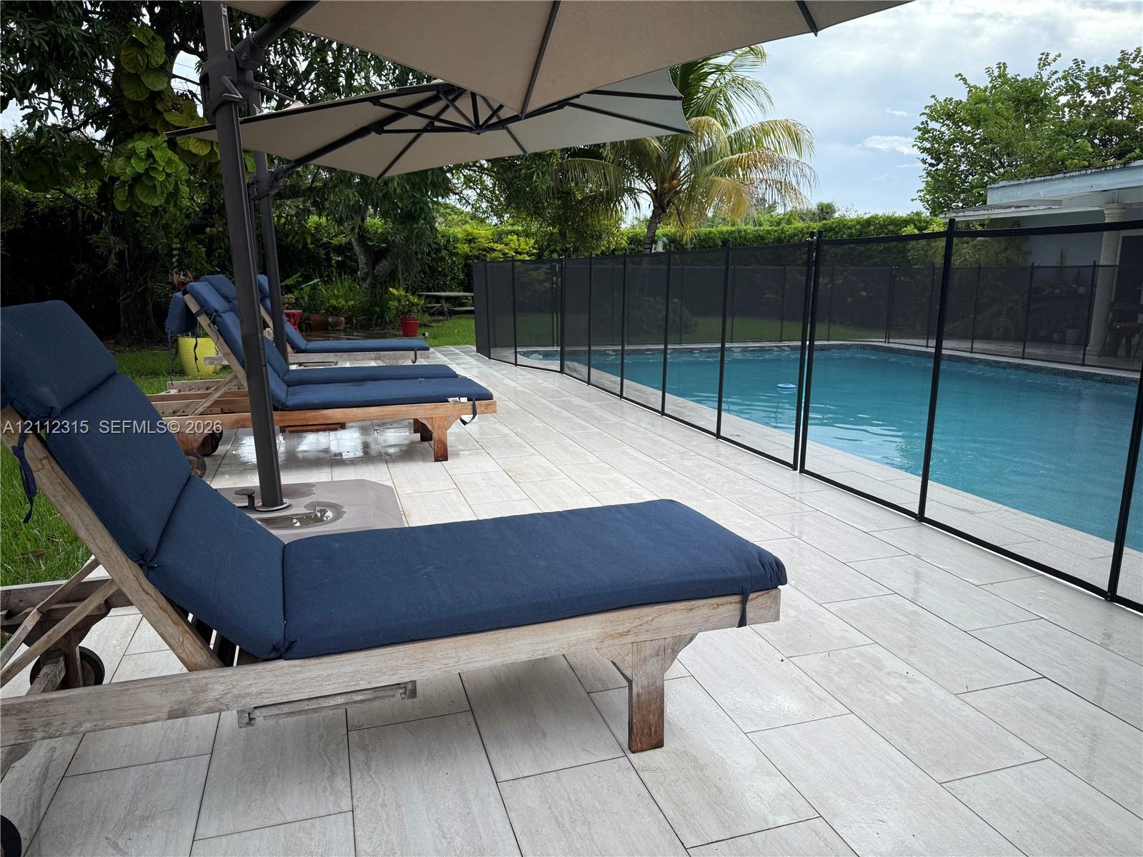 In-ground swimming pool featuring a removable mesh safety fence and light-toned tile pool deck surrounded by mature landscaping