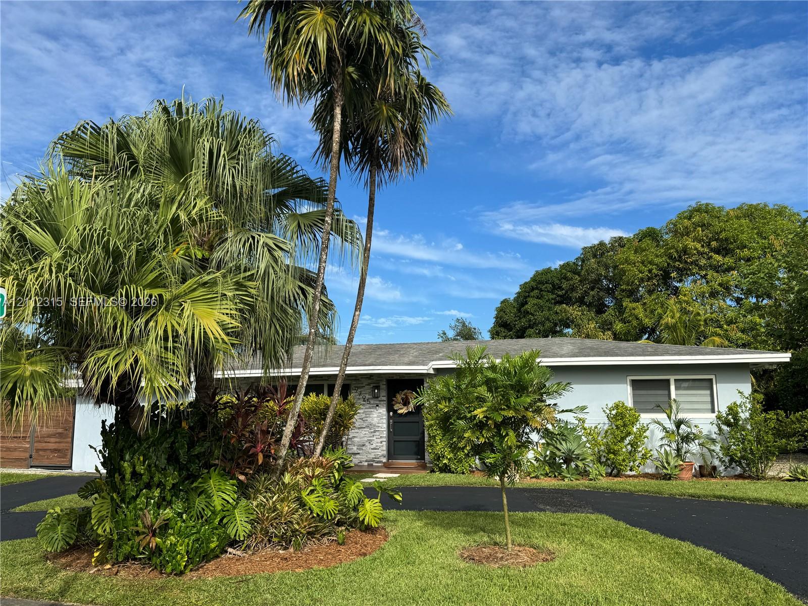 Single-story exterior featuring light-toned horizontal siding, a stone veneer facade, an asphalt driveway, and mature palm trees Single-story exterior featuring light-toned horizontal siding, a stone veneer facade, an asphalt driveway, and mature palm trees