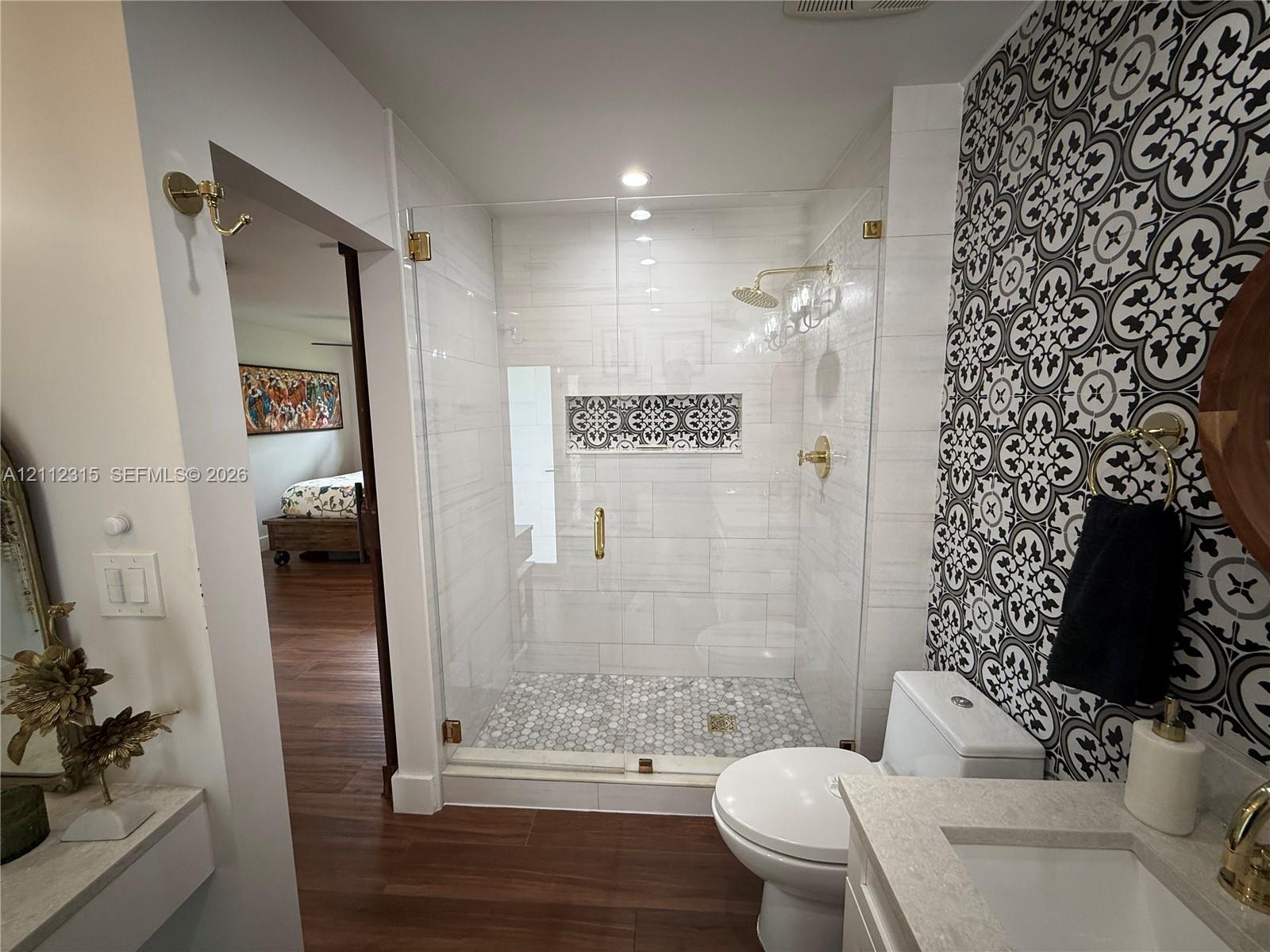 Walk-in shower featuring a glass enclosure, brass-finish fixtures, a decorative tile niche, and penny-round tile flooring, complemented by a patterned tile accent wall and stone-surfaced vanity