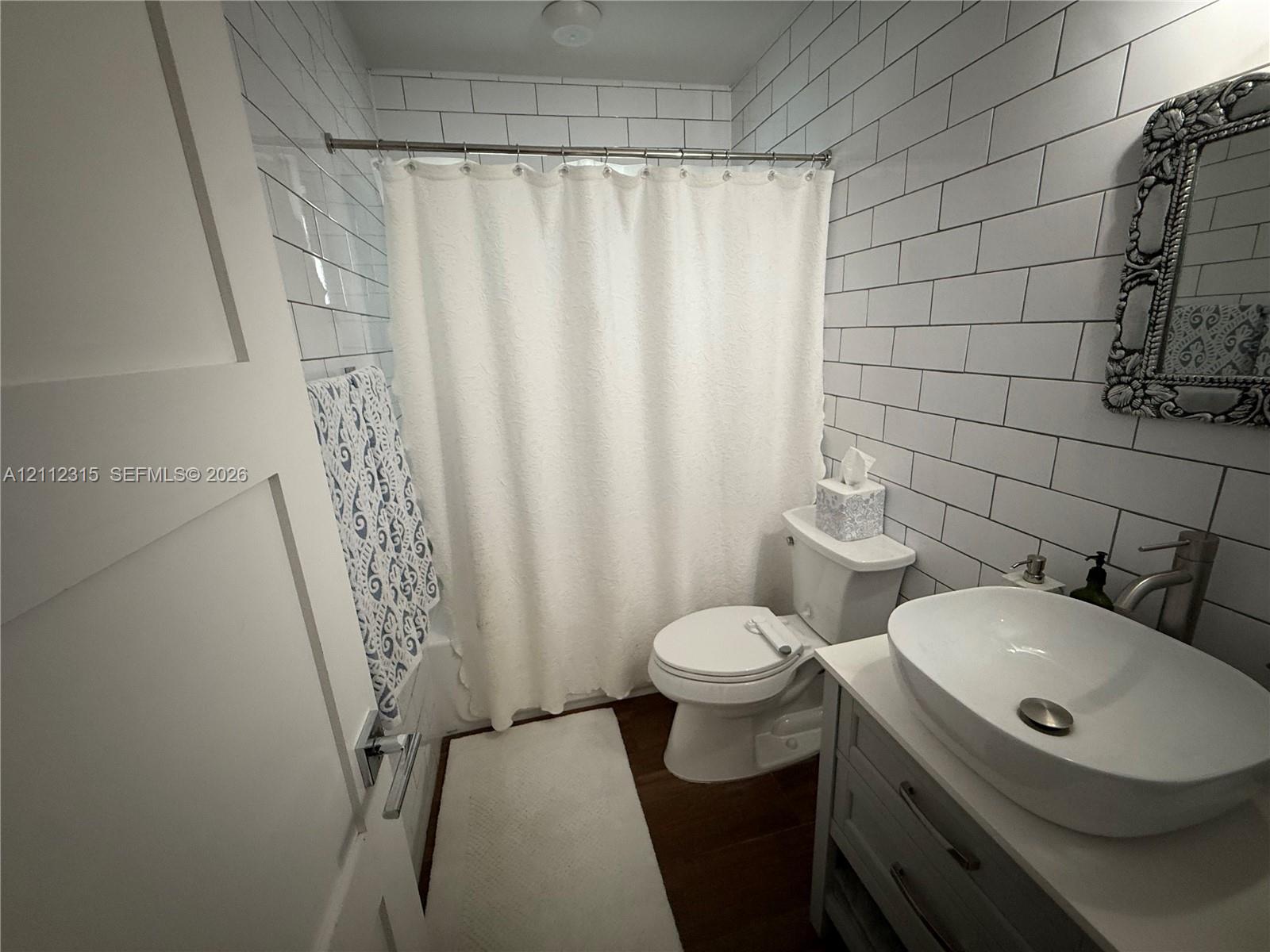 Bathroom featuring subway tile wall finishes, a white vanity cabinet, a rounded vessel sink with a metal faucet, and wood-finish flooring