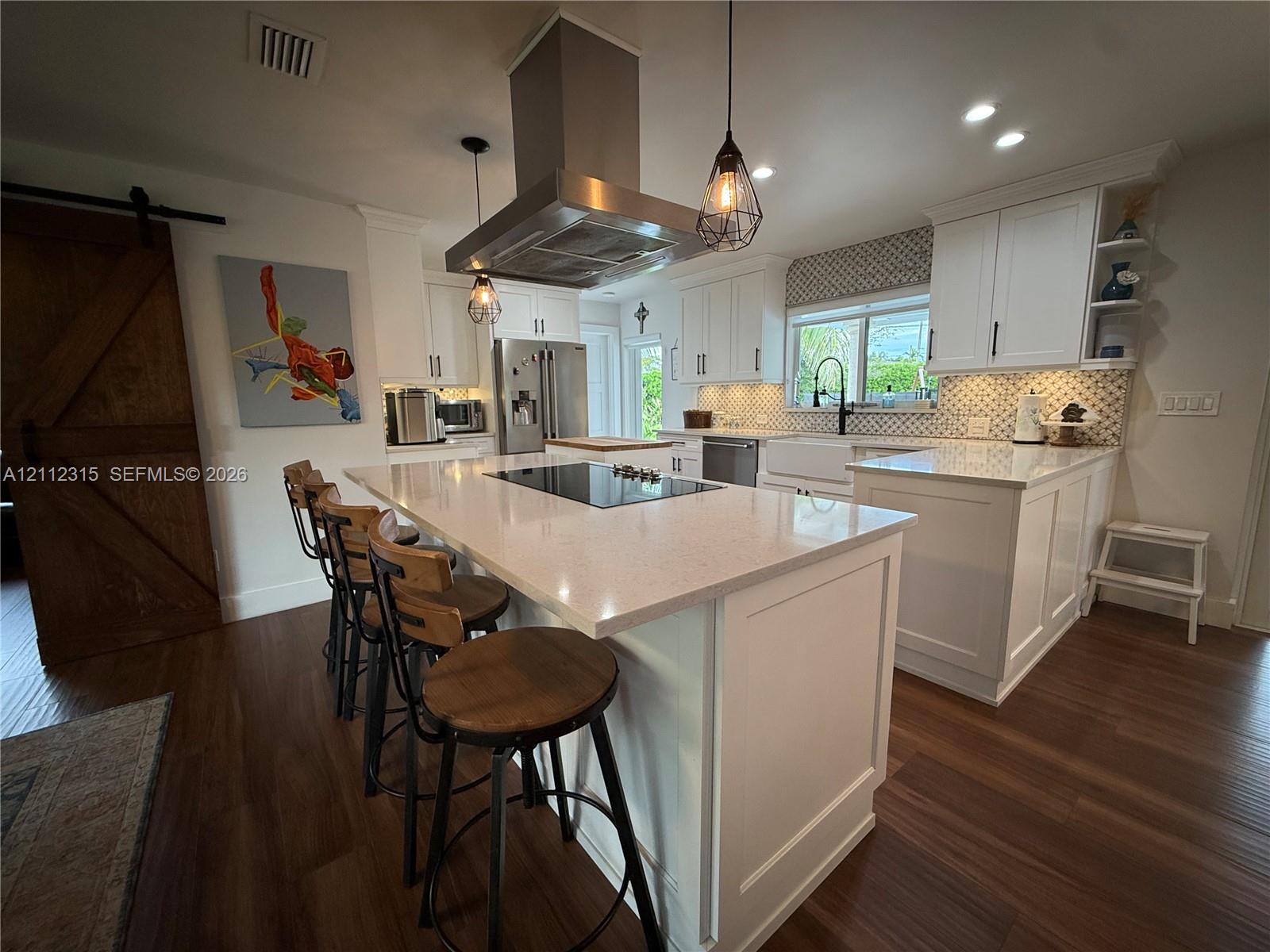Kitchen featuring white shaker-style cabinetry, an expansive central island with a solid-surface countertop and built-in cooktop, stainless steel vent hood, apron-front sink, and wood-finish flooring