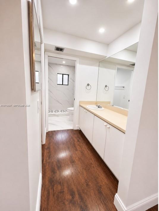 Bathroom featuring wood-finish flooring, white cabinetry, a solid-surface countertop with an integrated sink, full-width mirror, and recessed lighting