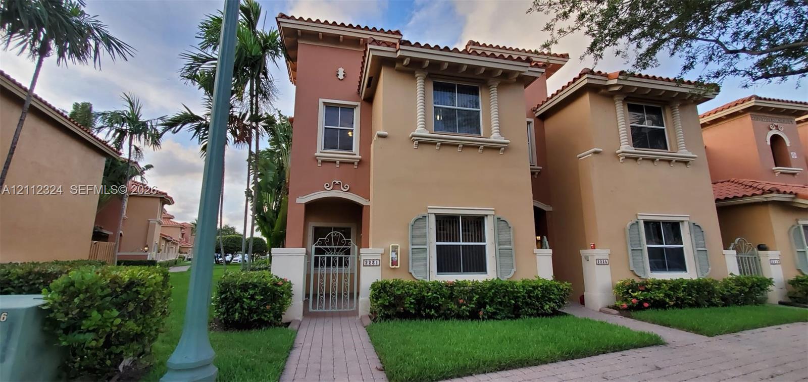 Two-story stucco exterior featuring a tile roof, decorative corbels, louvered shutters, and a gated entrance