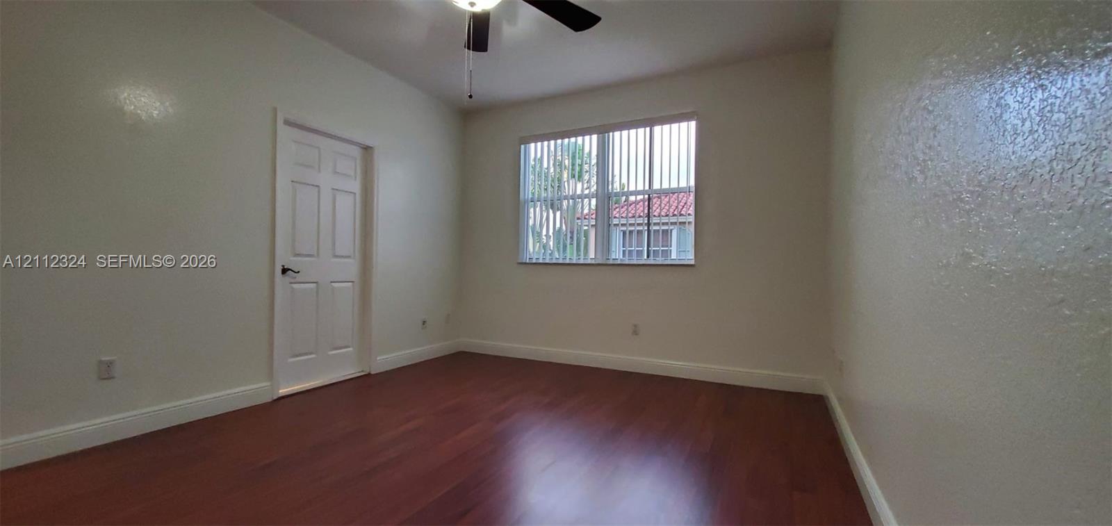 Interior room featuring wood-finish flooring, a multi-pane window with vertical blinds, a six-panel interior door, and a ceiling-mounted light fixture with fan