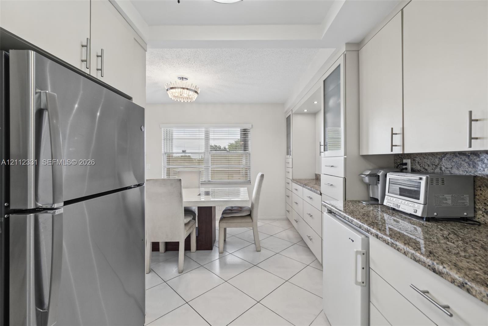 Kitchen featuring white cabinetry with brushed metal hardware, speckled stone countertops, stainless steel refrigerator, white ceramic tile flooring, and a crystal-accented chandelier
