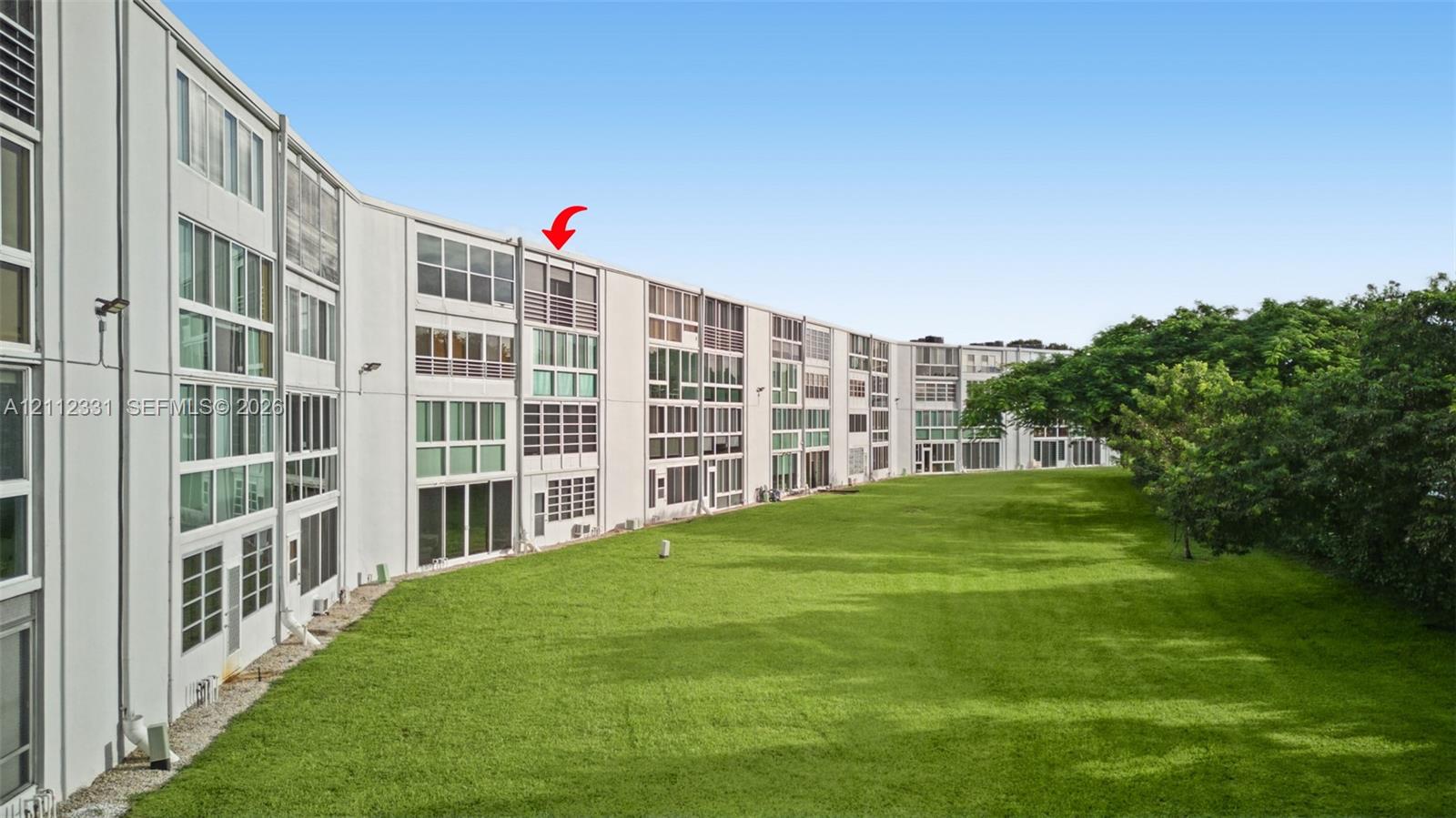 Multi-story exterior featuring a grid-pattern facade, extensive floor-to-ceiling glass windows, integrated privacy screens, and an expansive lawn bordered by mature trees