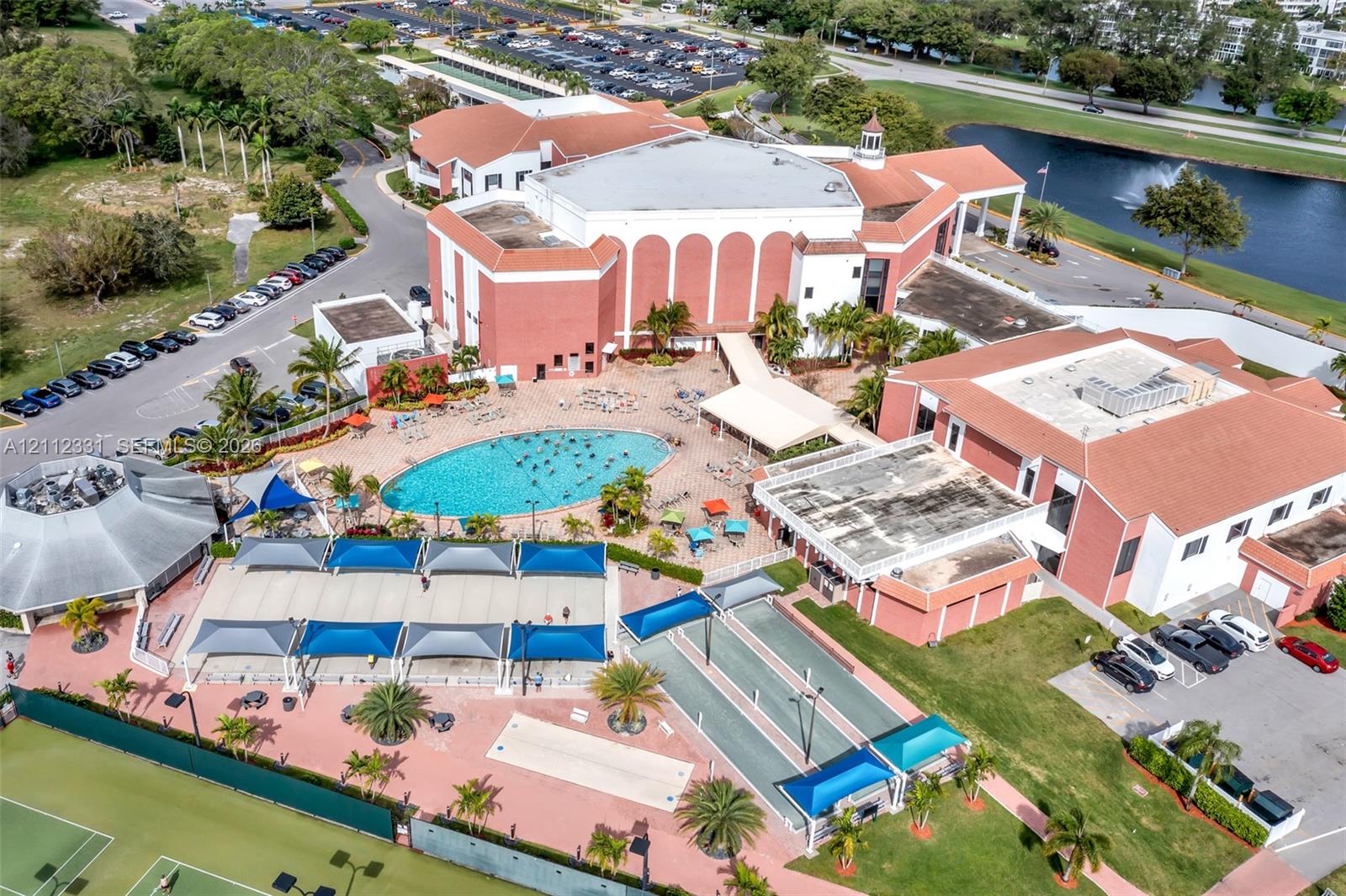 Aerial perspective of the property featuring a swimming pool, multiple tennis courts, shaded patio structures, and dedicated shuffleboard areas